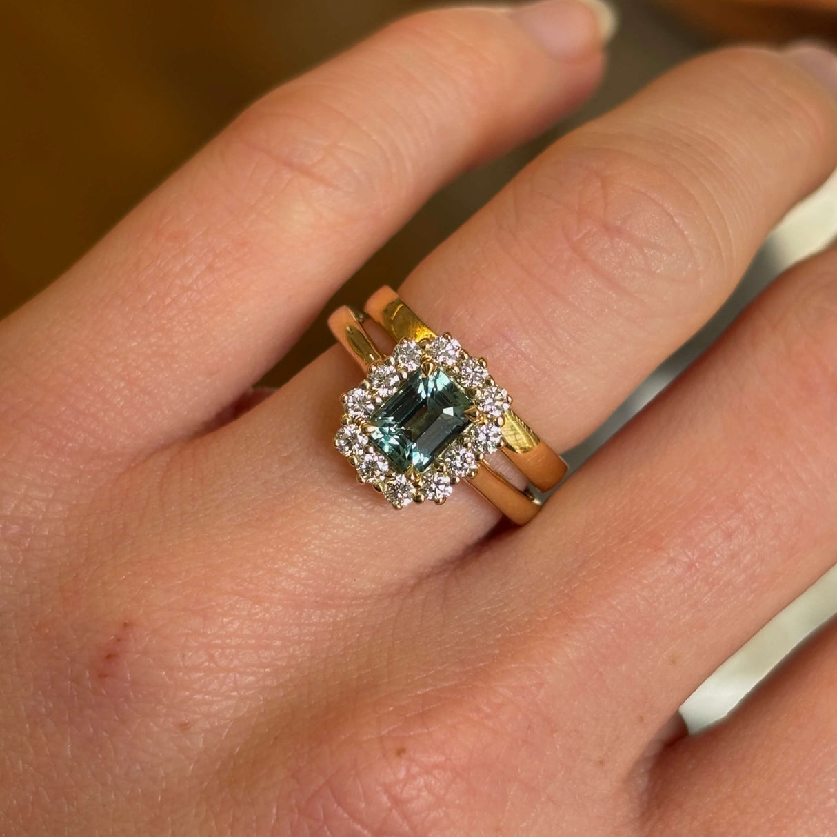 A yellow gold green sapphire and diamond cluster engagement ring worn on hand paired with a wedding band to show scale.