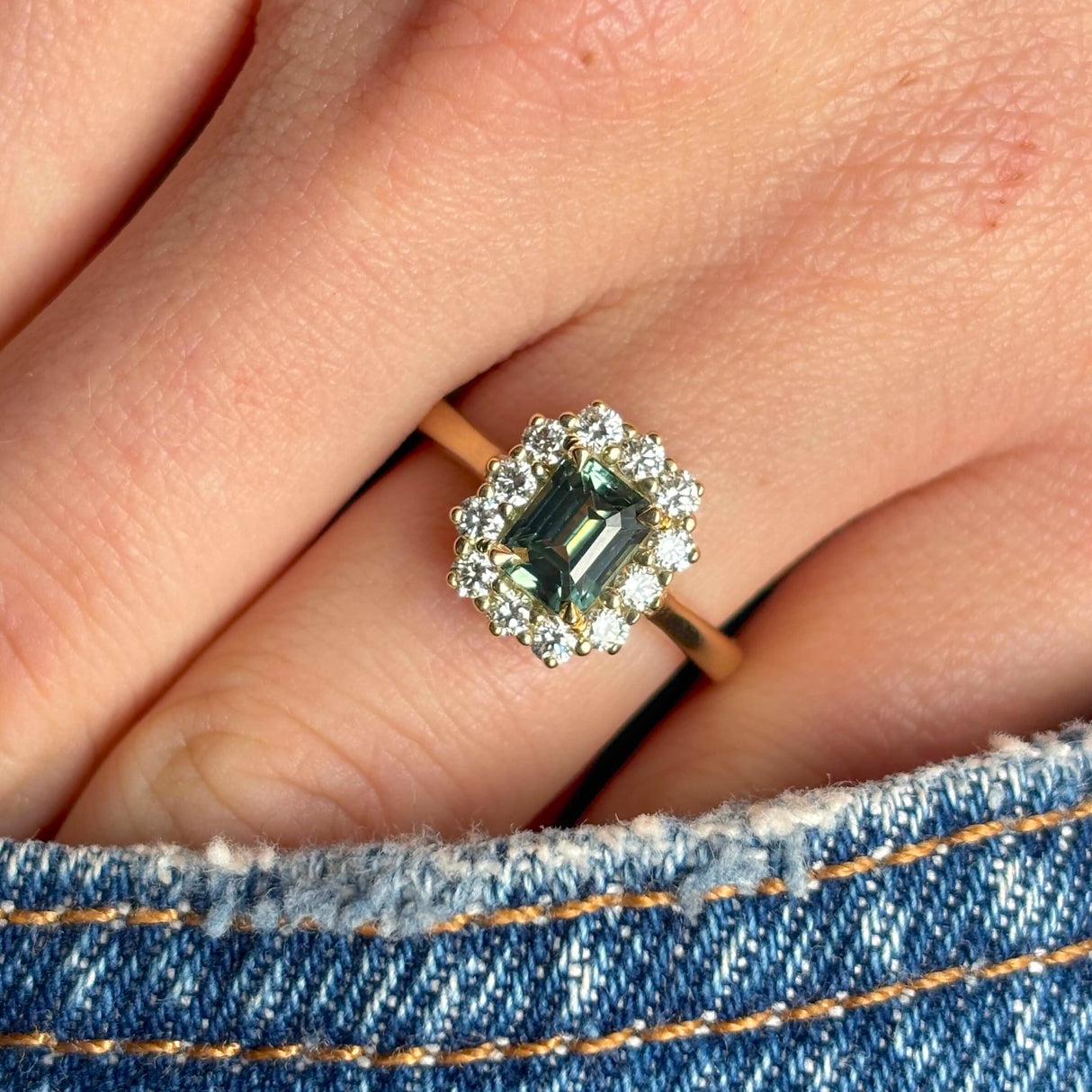 A yellow gold green sapphire and diamond cluster engagement ring worn on hand, in pocket close up to show the colour of the gemstone.