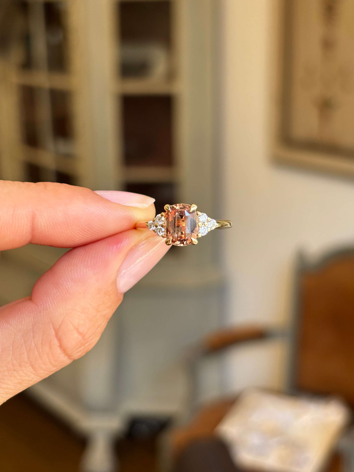 A close-up of a hand holding a vintage-inspired gold engagement ring with a central peach-colored rectangular sapphire flanked by diamonds, set against a softly blurred indoor background.