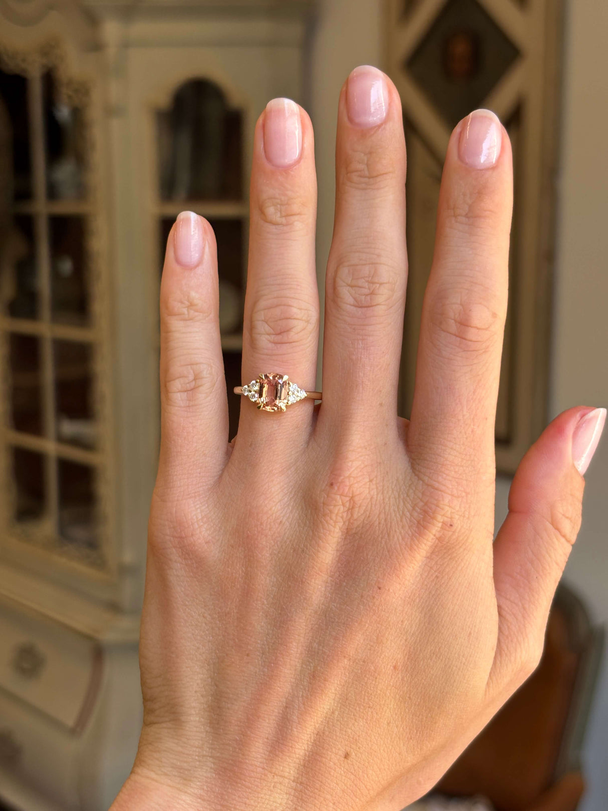 A Vintage-inspired yellow gold engagement ring worn on a person's ring finger, shown in a close-up hand shot with a softly blurred indoor background. Showcasing the central orange sapphire and diamonds.