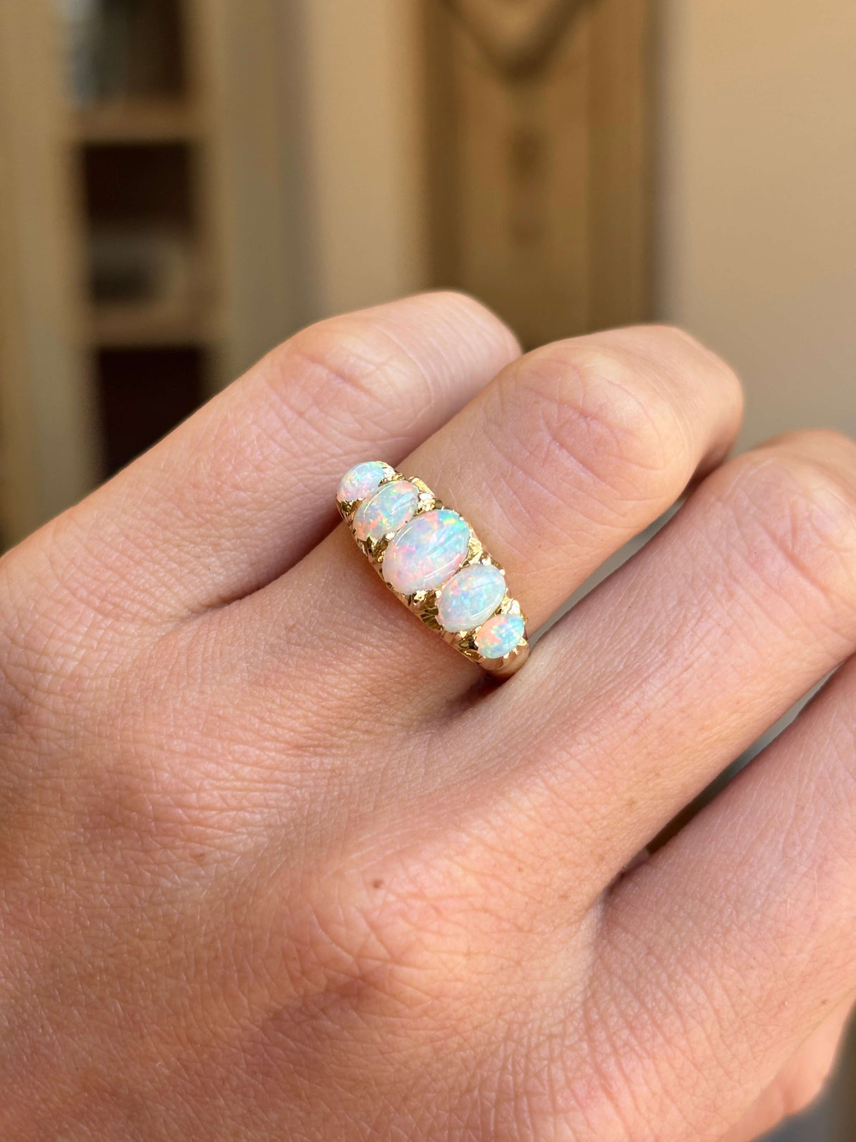 An Antique, Victorian yellow gold ring featuring five opal gemstones in a row, worn on a person's finger. The opal stones exhibit a milky white color with hints of blue and green.