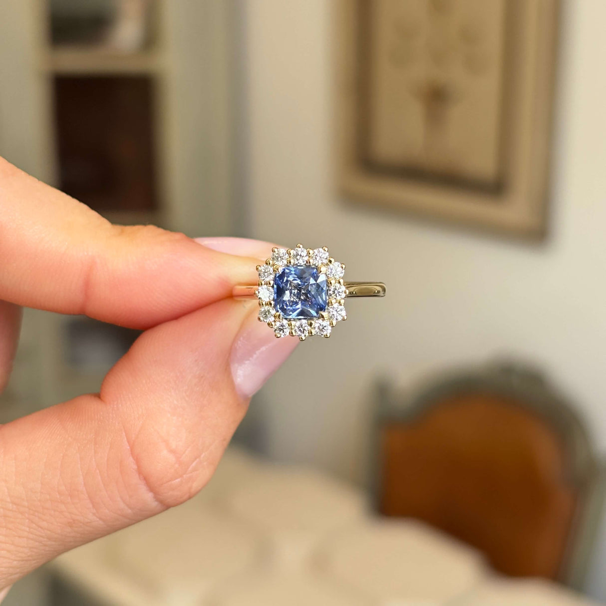 A hand holding an antique-inspired gold ring with a central blue sapphire and a halo of diamonds, set against a softly blurred indoor background.