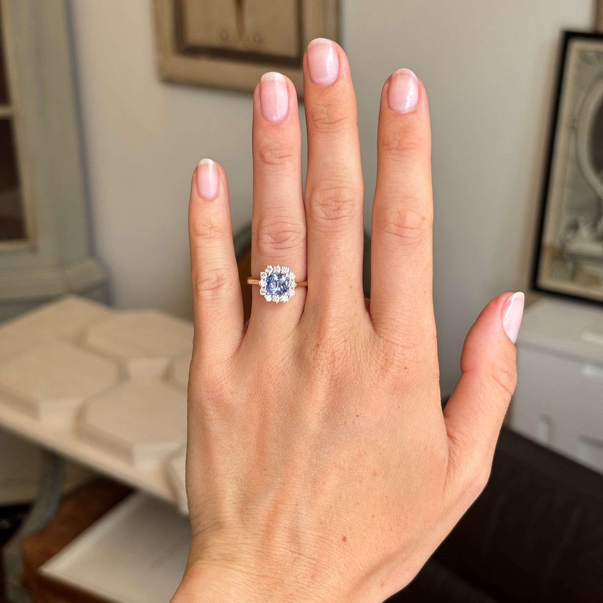 A close-up of a hand wearing an antique-inspired yellow gold engagement ring set with a blue cushion-cut sapphire and a halo of diamonds, against a softly blurred indoor background.
