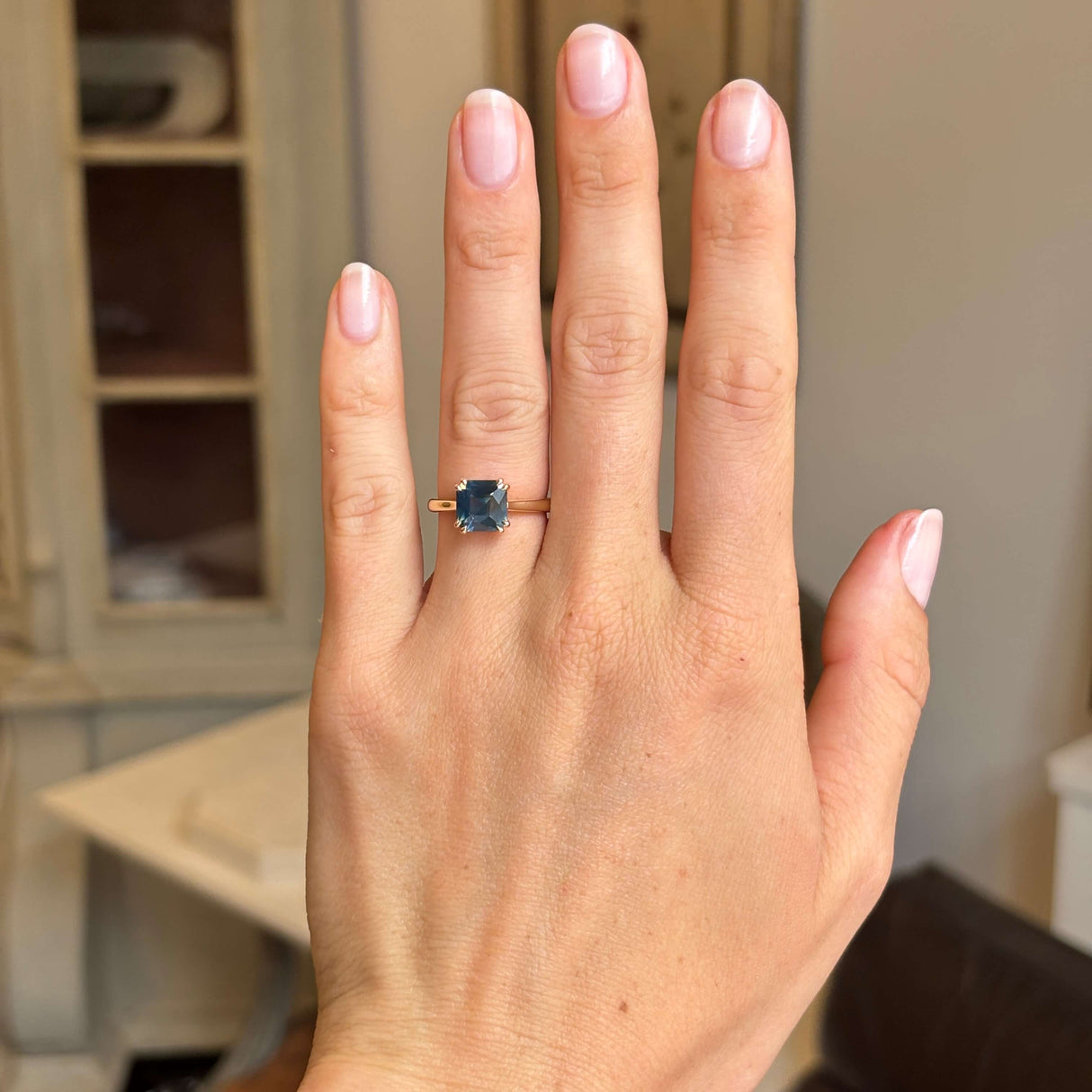 A close-up of a hand wearing an Antique-inspired yellow gold ring with a square-cut sapphire, set against a softly blurred indoor background.