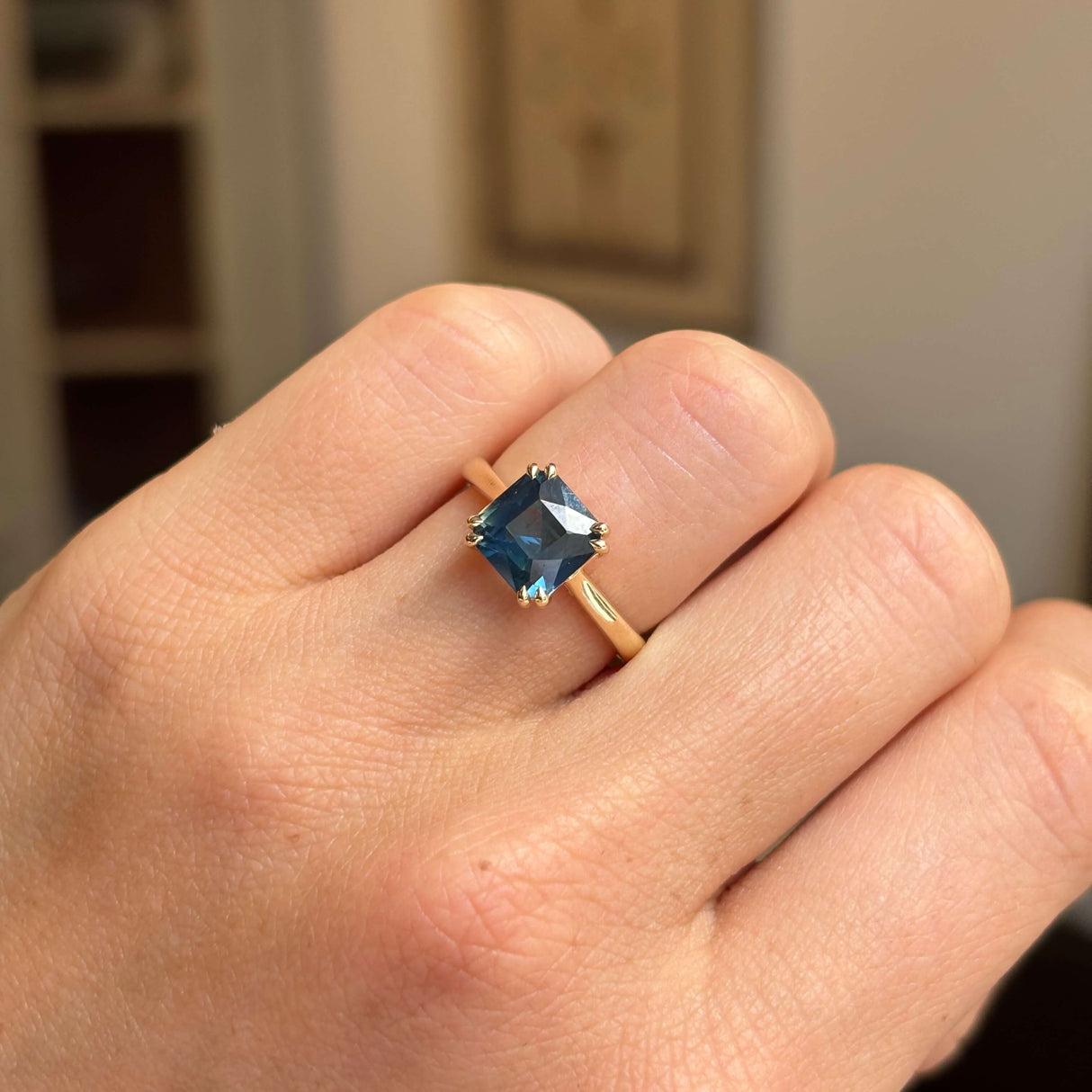 An Antique-inspired, teal square-cut sapphire solitaire ring worn on a person's finger, shown in a close-up shot against a softly blurred indoor background.