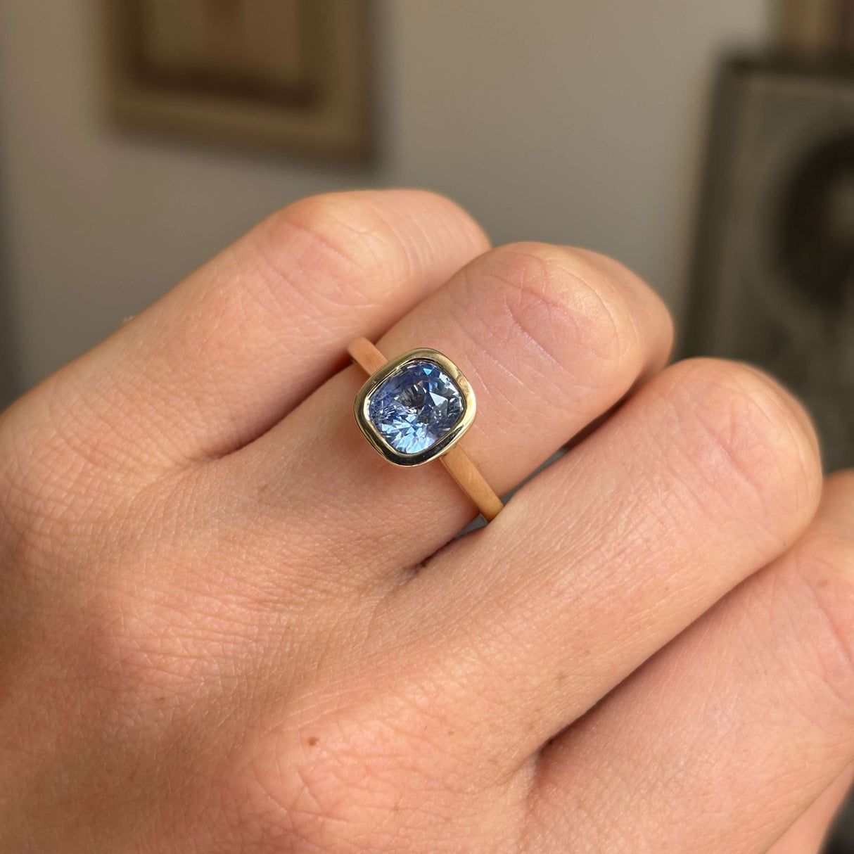 A close-up of a hand wearing a ring with a square cushion-cut blue gemstone in a gold bezel setting on a tan band, photographed indoors with a blurred background.