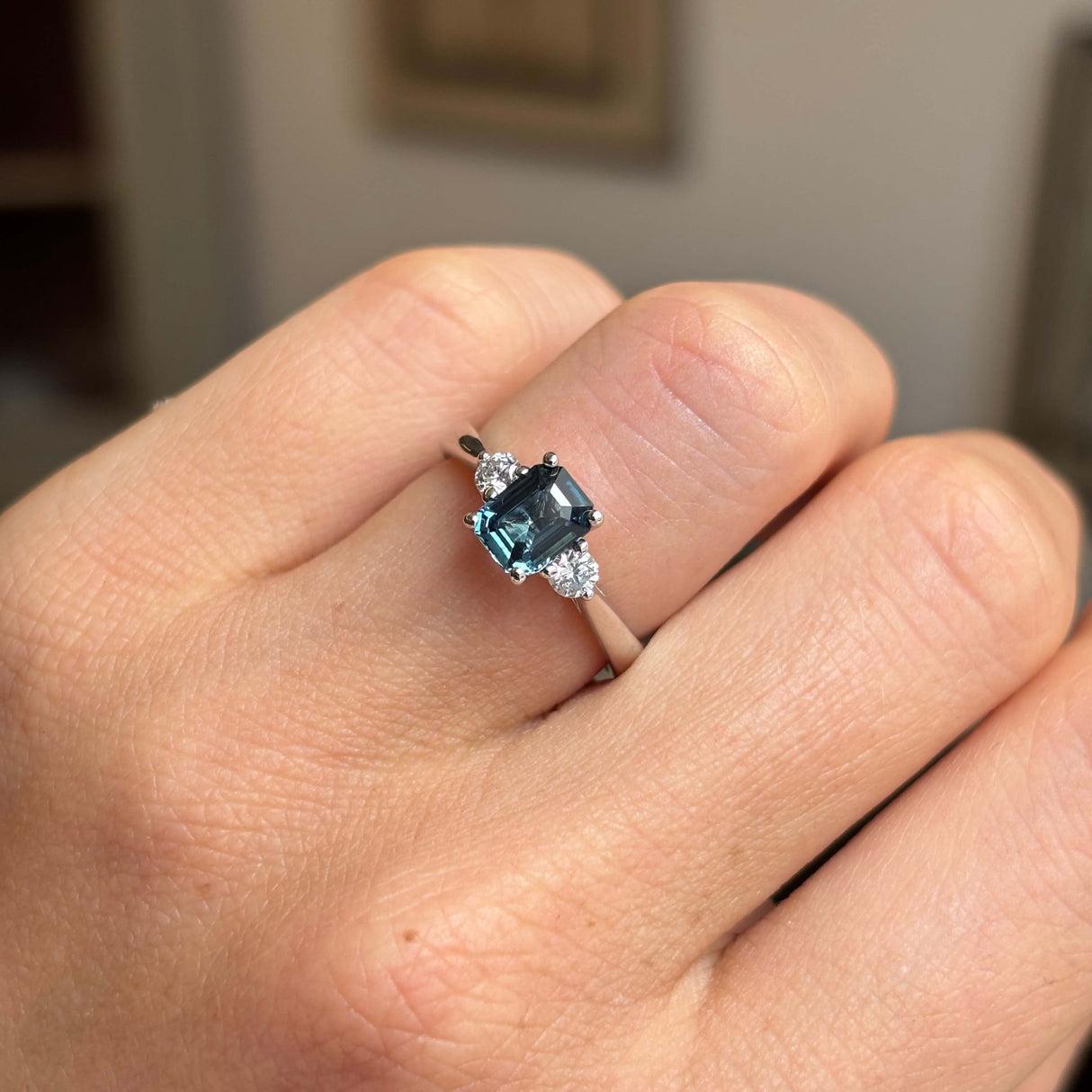 Vintage-inspired, Teal sapphire and diamond three-stone engagement ring in platinum on a person's finger against a neutral background