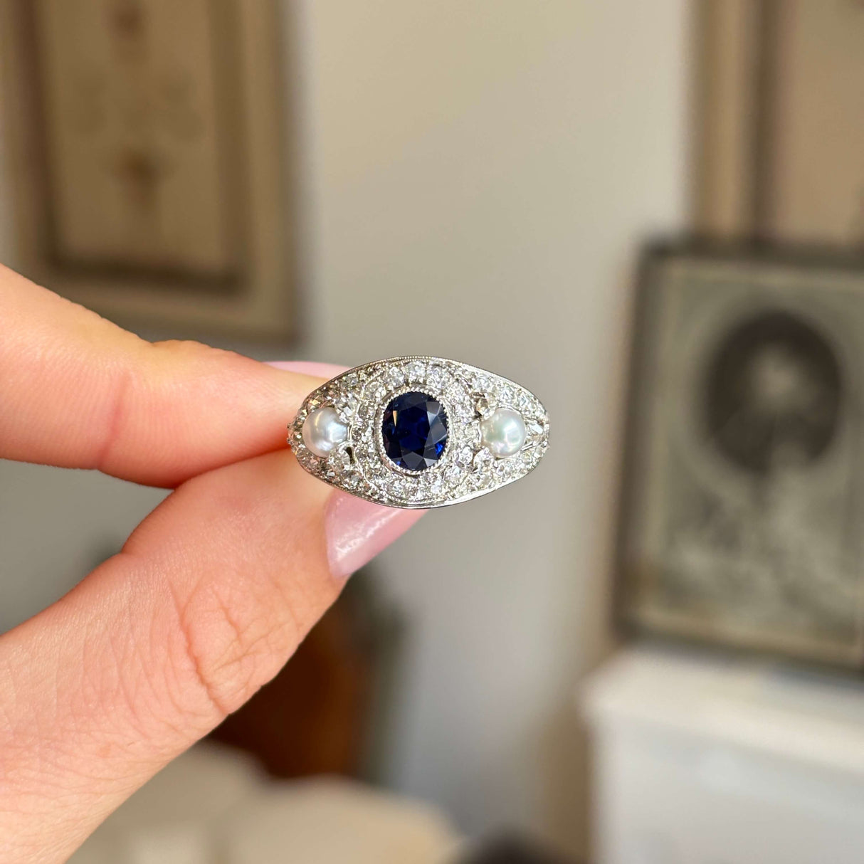 An Antique, Belle Époque dark blue sapphire, pearl and diamond cluster cocktail ring in white gold held by a hand against a blurred background
