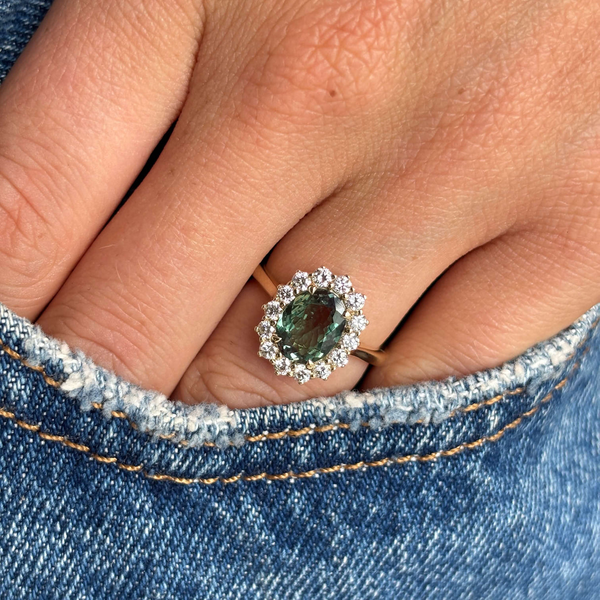 Close-up of a hand wearing an Antique-inspired, alexandrite and diamond cluster engagement ring in gold on denim fabric