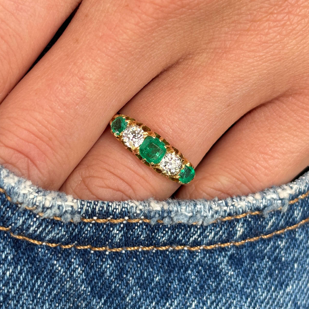 Close up of an Antique, Victorian gold five-stone ring with three old-mine emeralds and two old-mine cut diamonds worn on a finger against a denim background