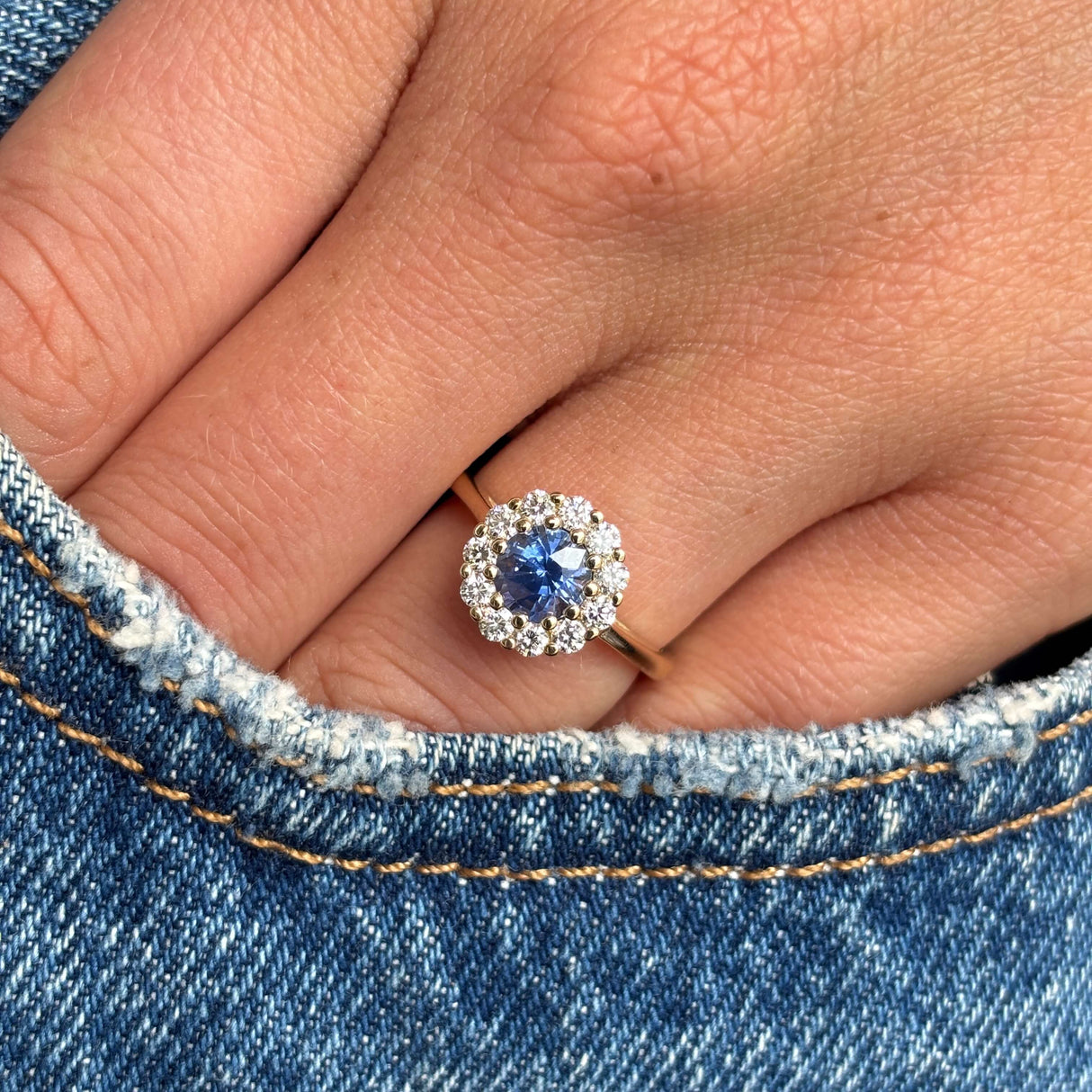 Close-up of a hand wearing an Antique-inspired, electric blue sapphire and diamond cluster engagement ring in yellow gold on denim fabric.