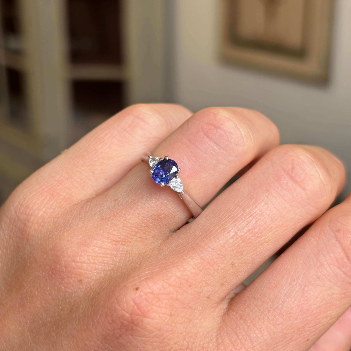 Hand wearing a Vintage-inspired, purple sapphire & diamond three-stone platinum engagement ring on a blurred indoor background
