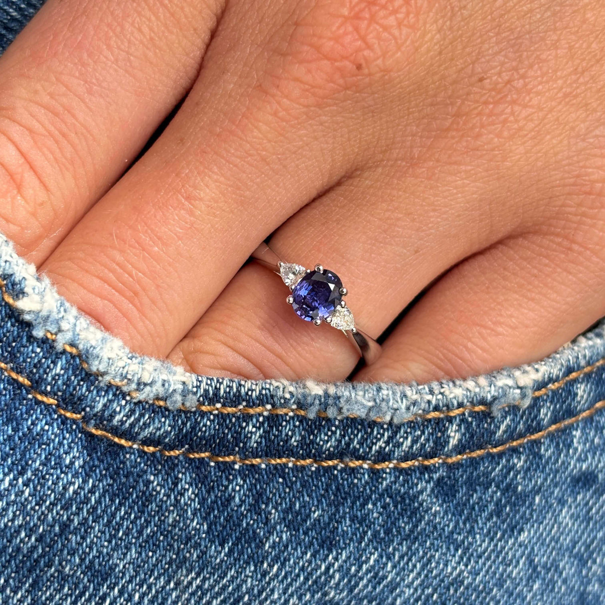 Hand wearing a platinum Vintage-inspired, deep blue sapphire & diamond three-stone engagement ring on denim fabric