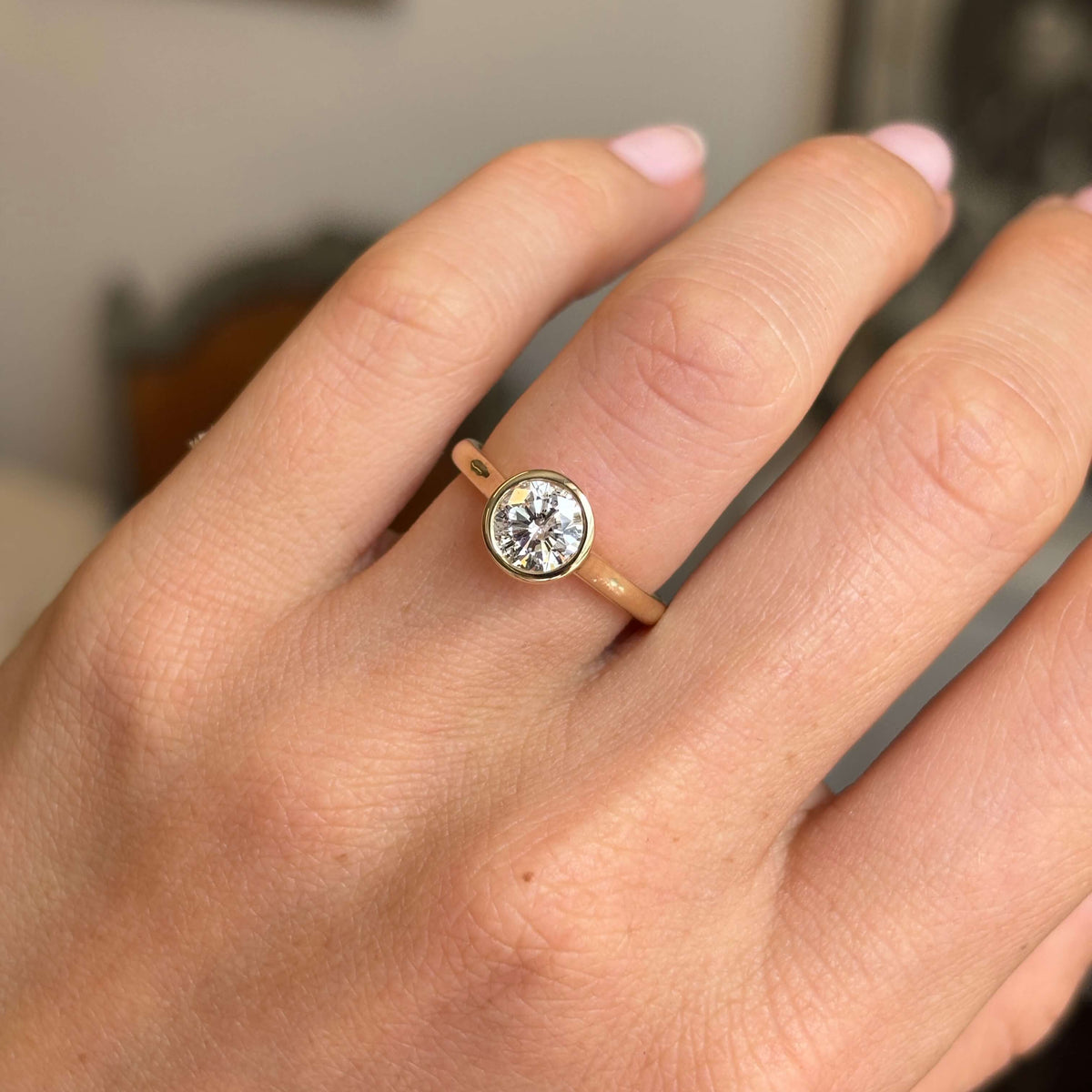 A close up of a 'salt and pepper' diamond in an antique-inspired solitaire engagement ring, worn on a hand showcasing the yellow gold rub-over setting and band, against a softly blurred background.