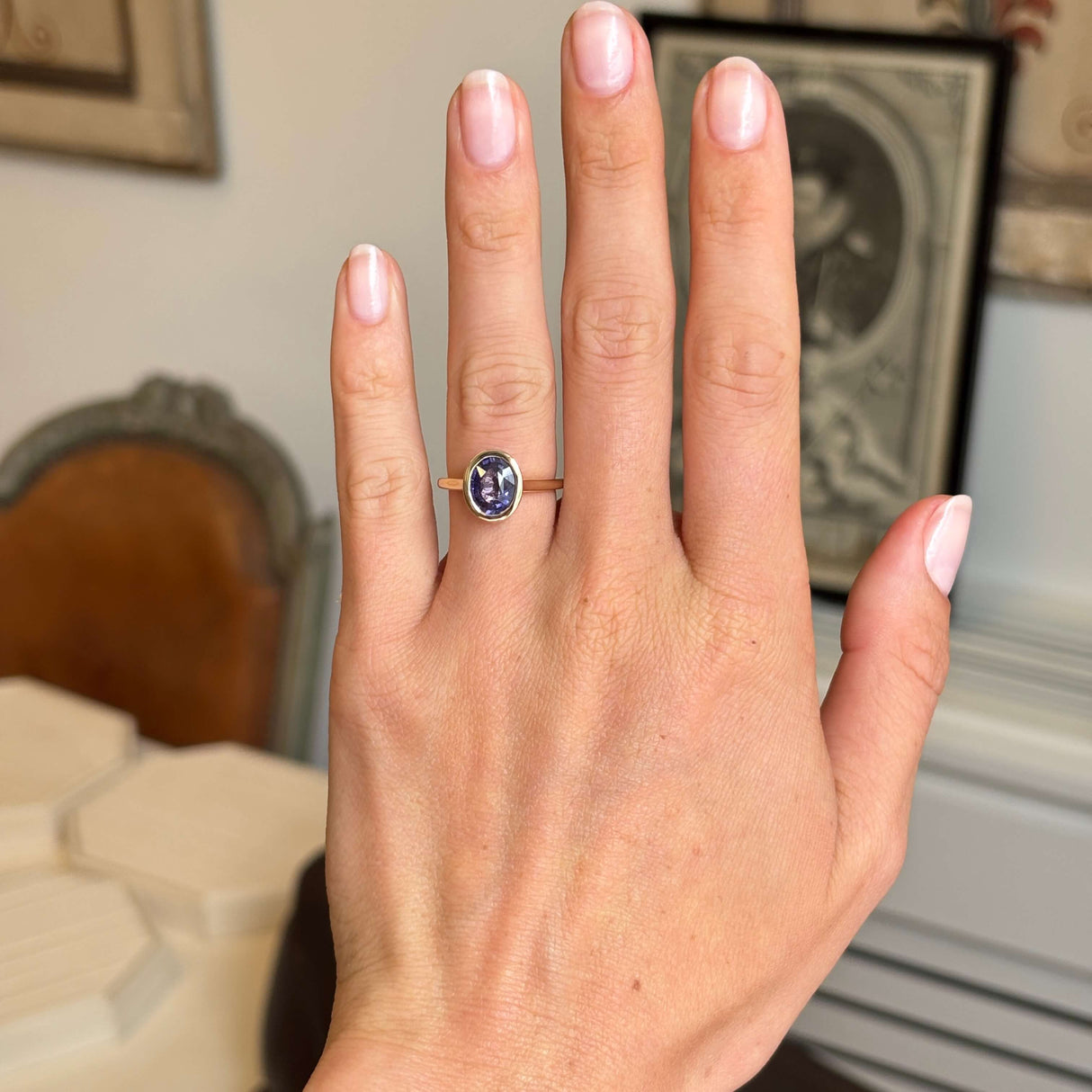 A hand wearing an Antique-inspired, purple oval sapphire solitaire engagement ring, set against a softly blurred indoor background.