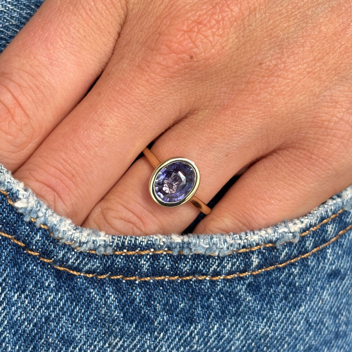 A close-up of a hand wearing a sapphire ring with an oval purple sapphire in a gold bezel setting, on a gold band, resting against blue denim jeans.