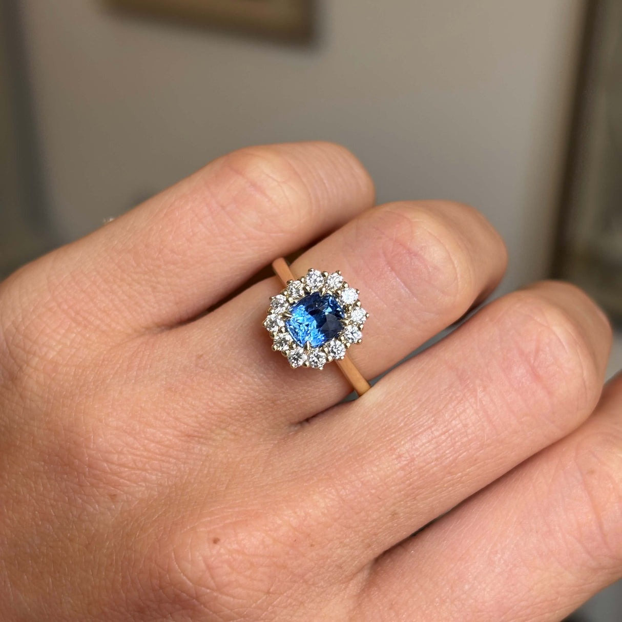 A close-up of a hand wearing an antique-inspired yellow gold ring with a central blue sapphire surrounded by a halo of diamonds.