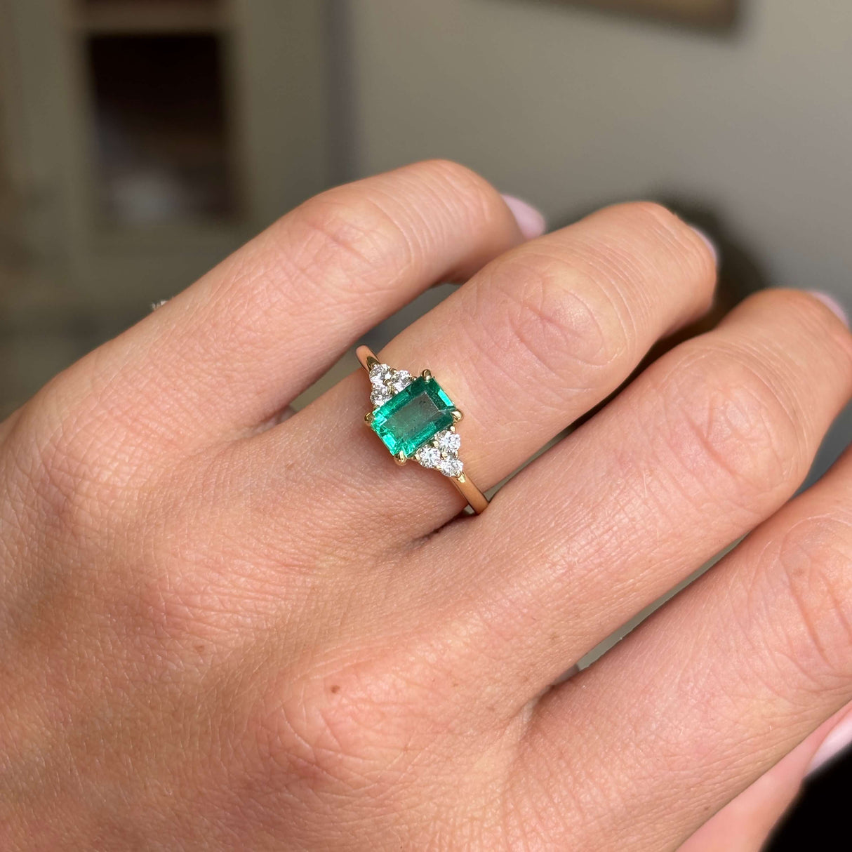 Hand wearing an Antique-inspired, Emerald and diamond cluster trefoil yellow gold engagement ring on a neutral background