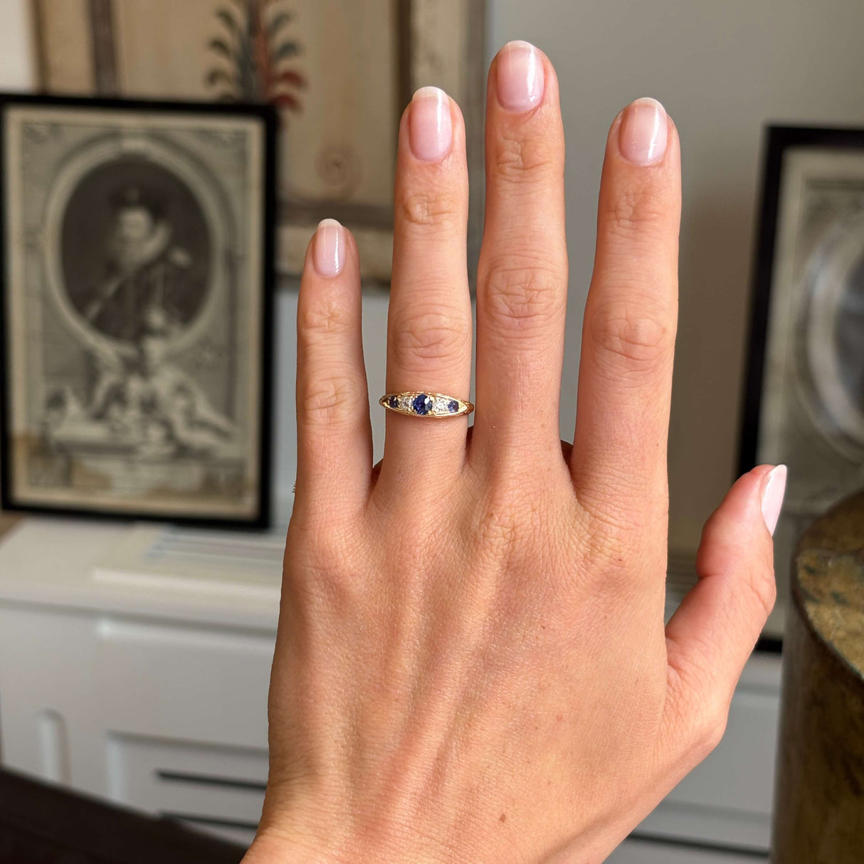 Hand wearing a Antique, Victorian sapphire & diamond five-stone engagement ring in yellow gold set against a blurred indoor background.