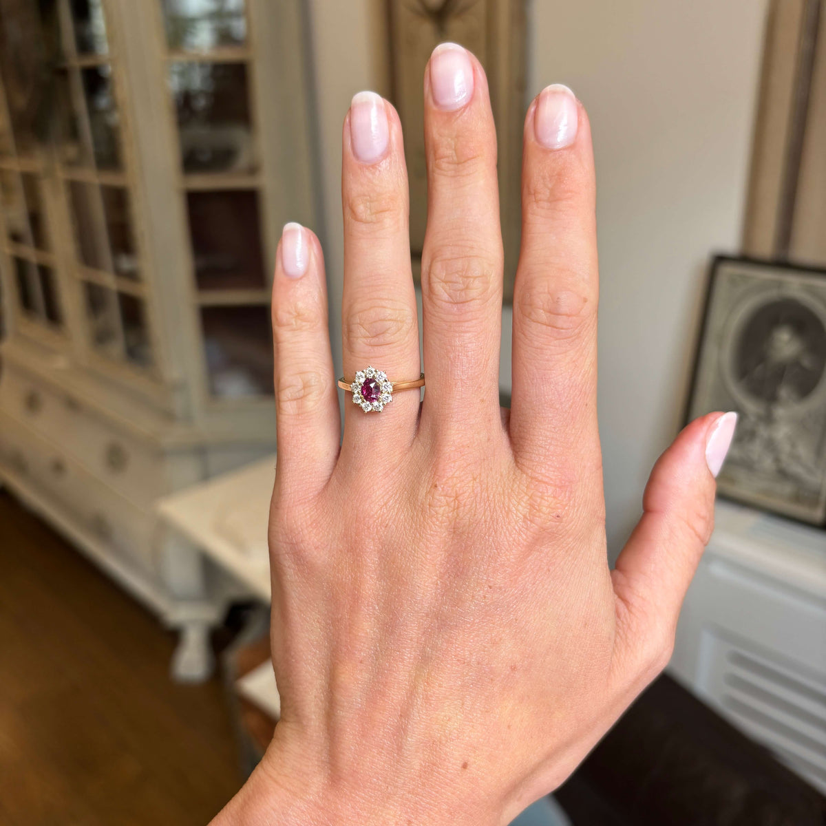 A close-up of a hand wearing an antique-inspired cluster engagement ring with a round pinkish-red central ruby surrounded by a halo of diamonds, on a gold band, photographed indoors with a blurred home background.