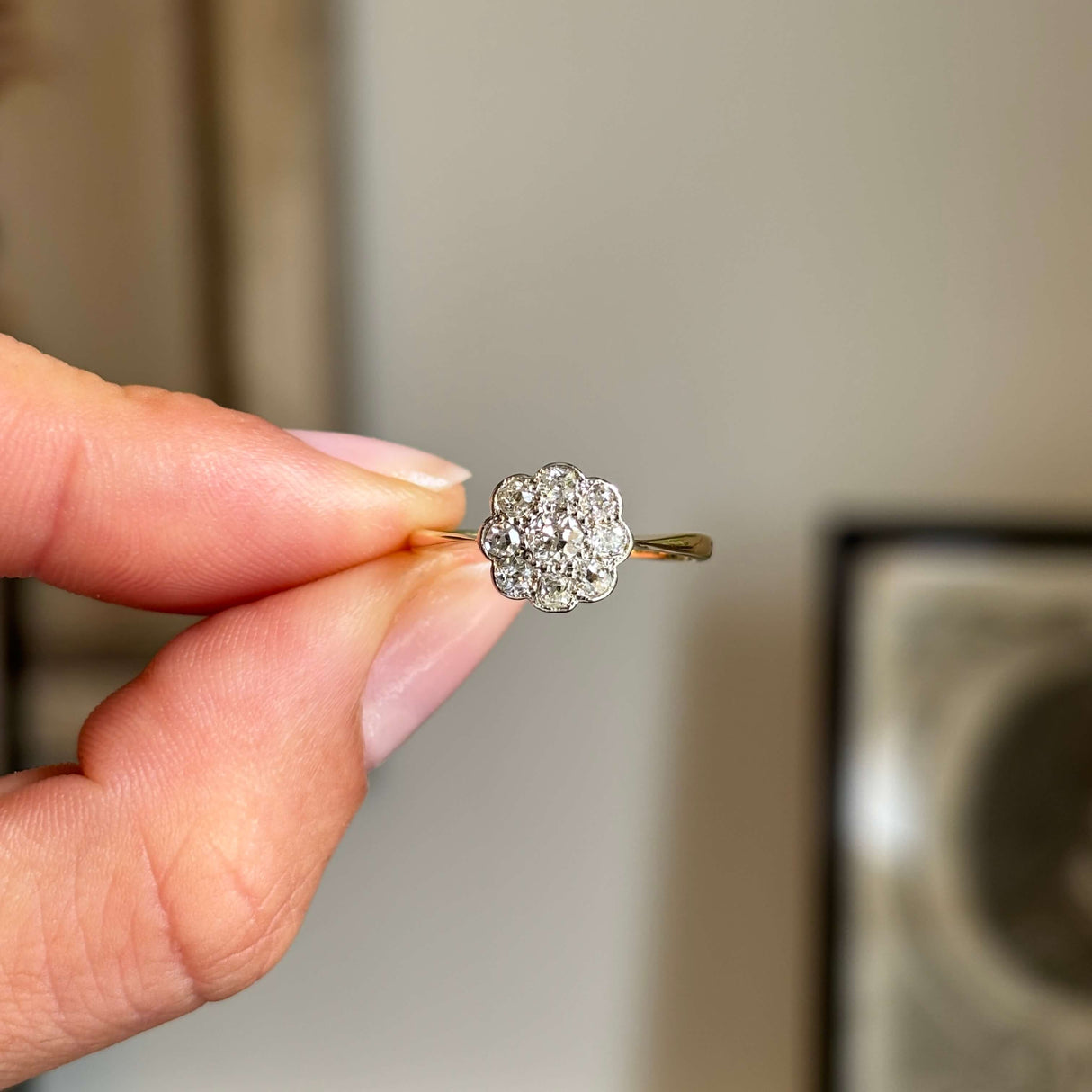 Hand holding a diamond daisy cluster engagement ring, from the Edwardian era, in yellow gold against a blurred background