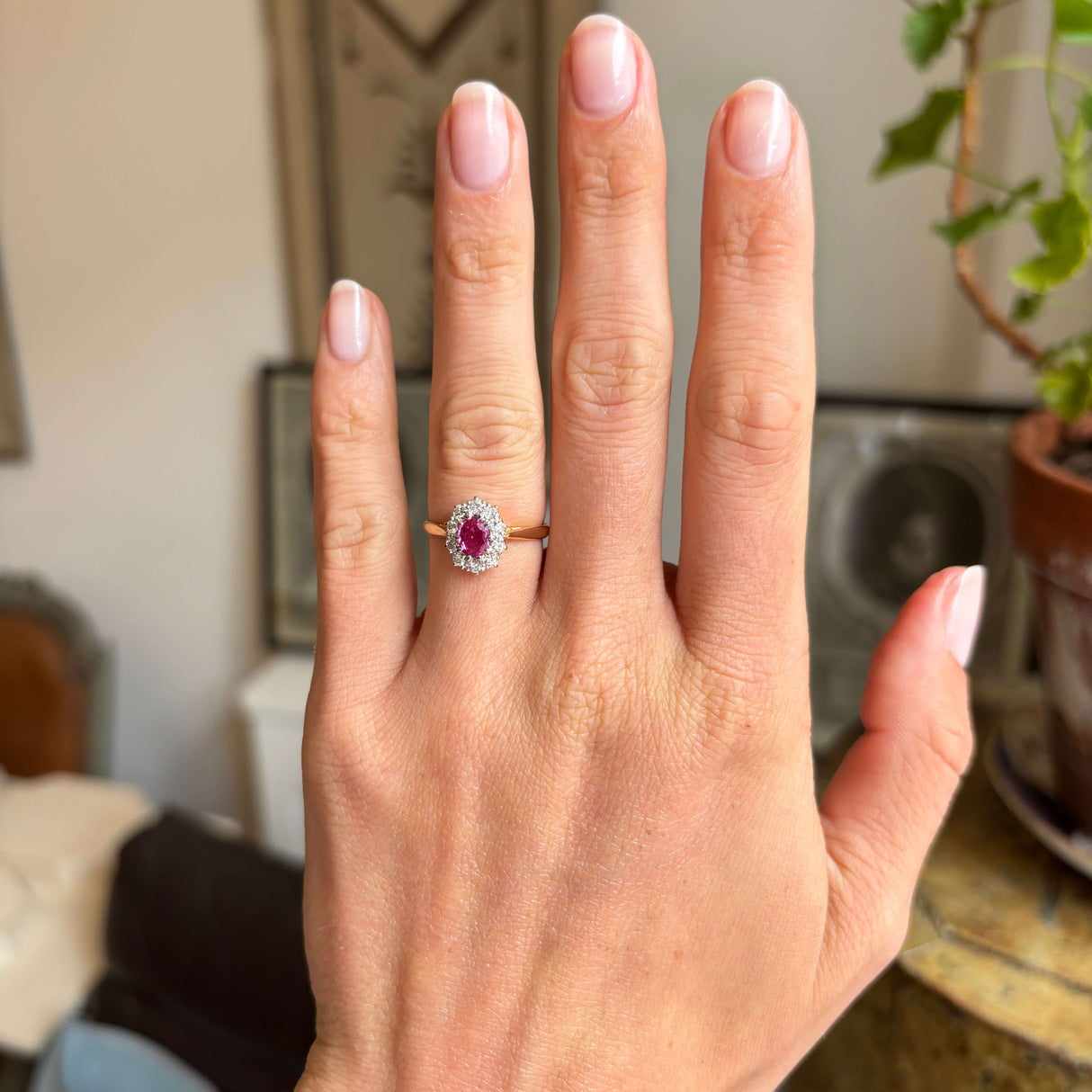 Hand wearing a Vintage 1940s yellow gold ring with a pink ruby at the centre and a diamond halo, blurred indoor background