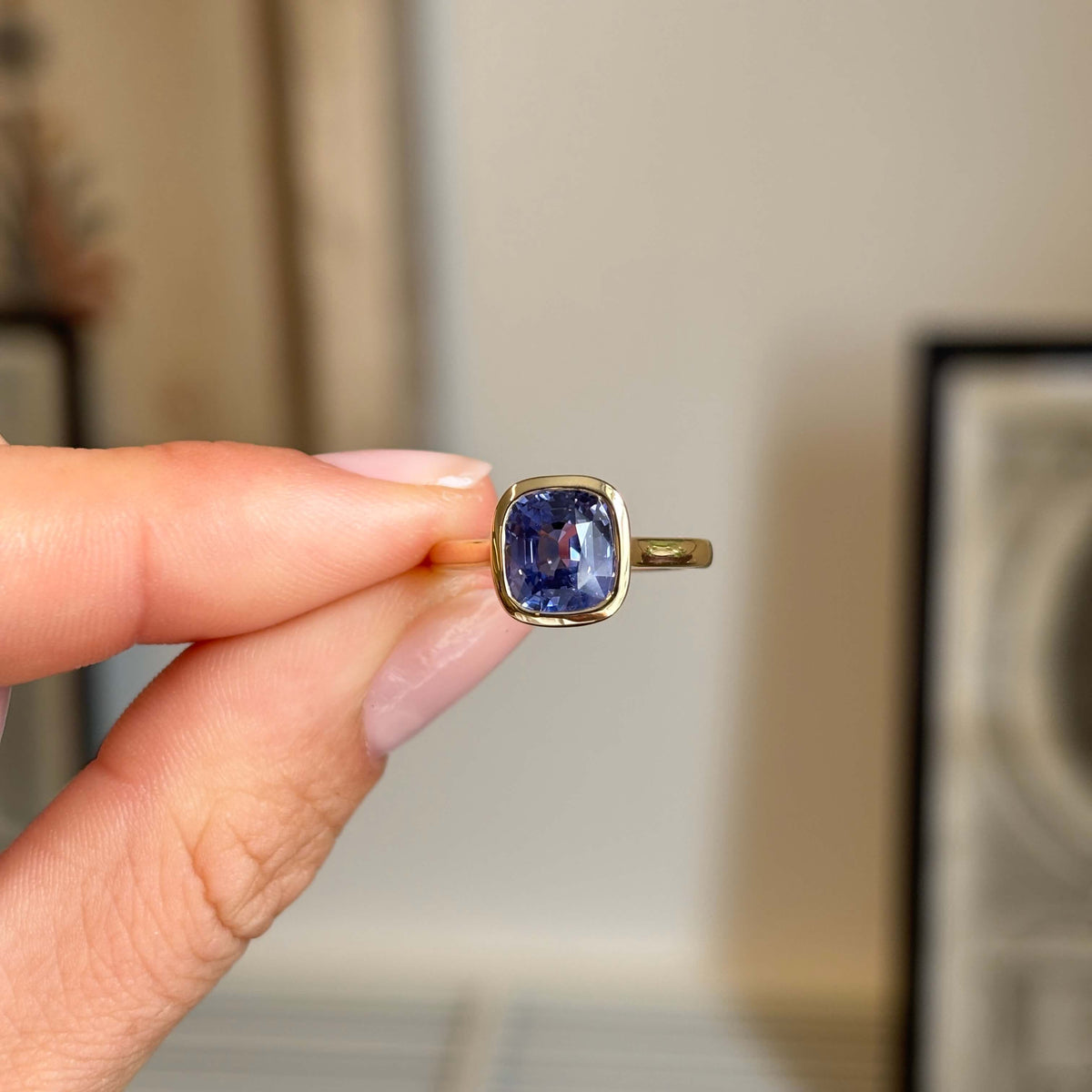 A gold ring with a cushion-cut purple sapphire gemstone held between fingers against a softly blurred indoor background. Antique-inspired