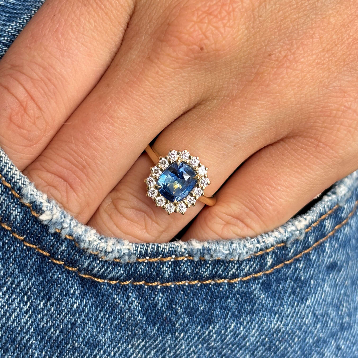 A close-up of a hand wearing a Antique-inspired, Blue cushion-cut sapphire and diamond cluster engagement ring in yellow gold, resting on blue denim.