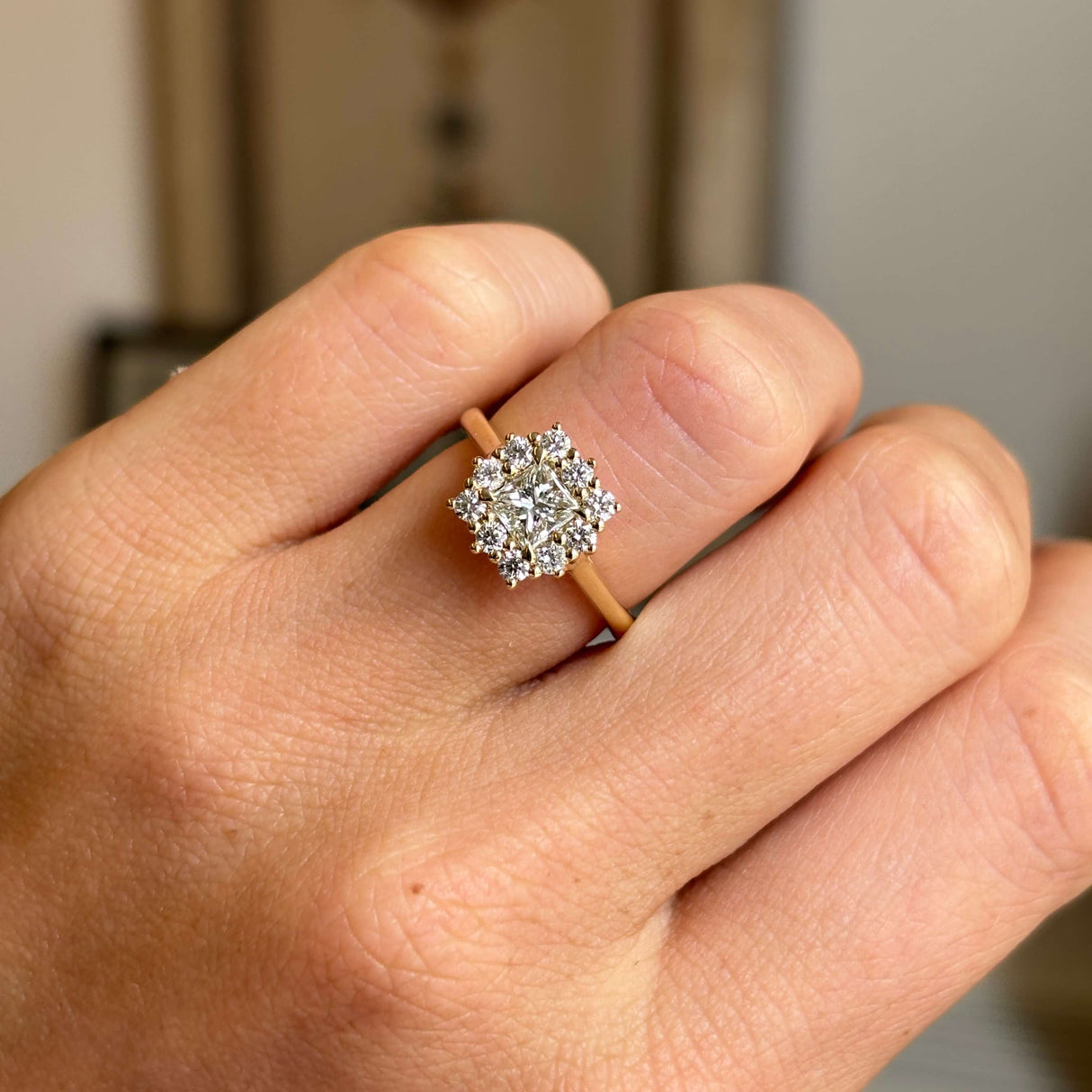 Hand wearing an Antique-inspired, princess-cut diamond cluster engagement ring in yellow gold on a blurred background