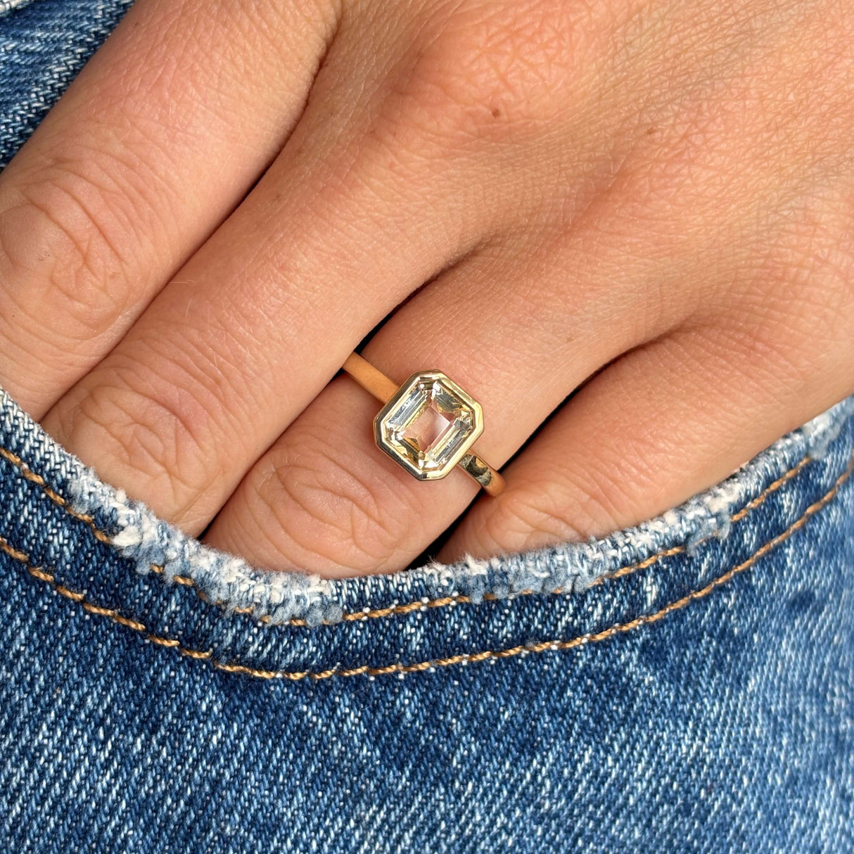Close-up of a hand wearing a gold ring with a pale yellow sapphire, set against a denim background. Antique-inspired.