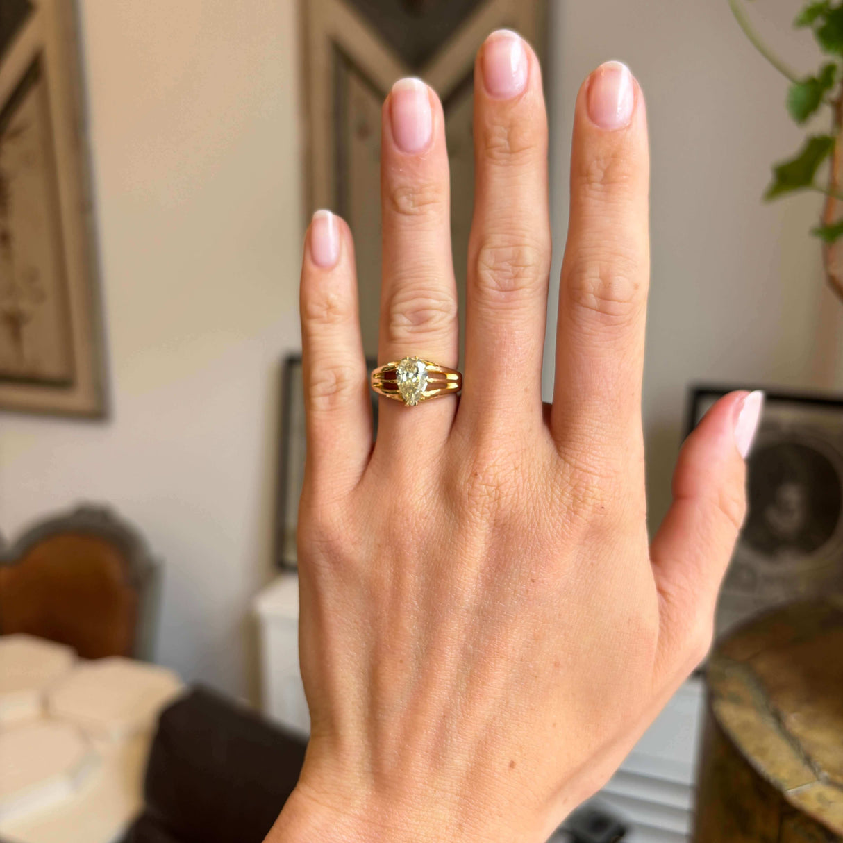 Hand wearing a gold Antique, Edwardian yellow diamond solitaire gypsy, set against a blurred indoor background.