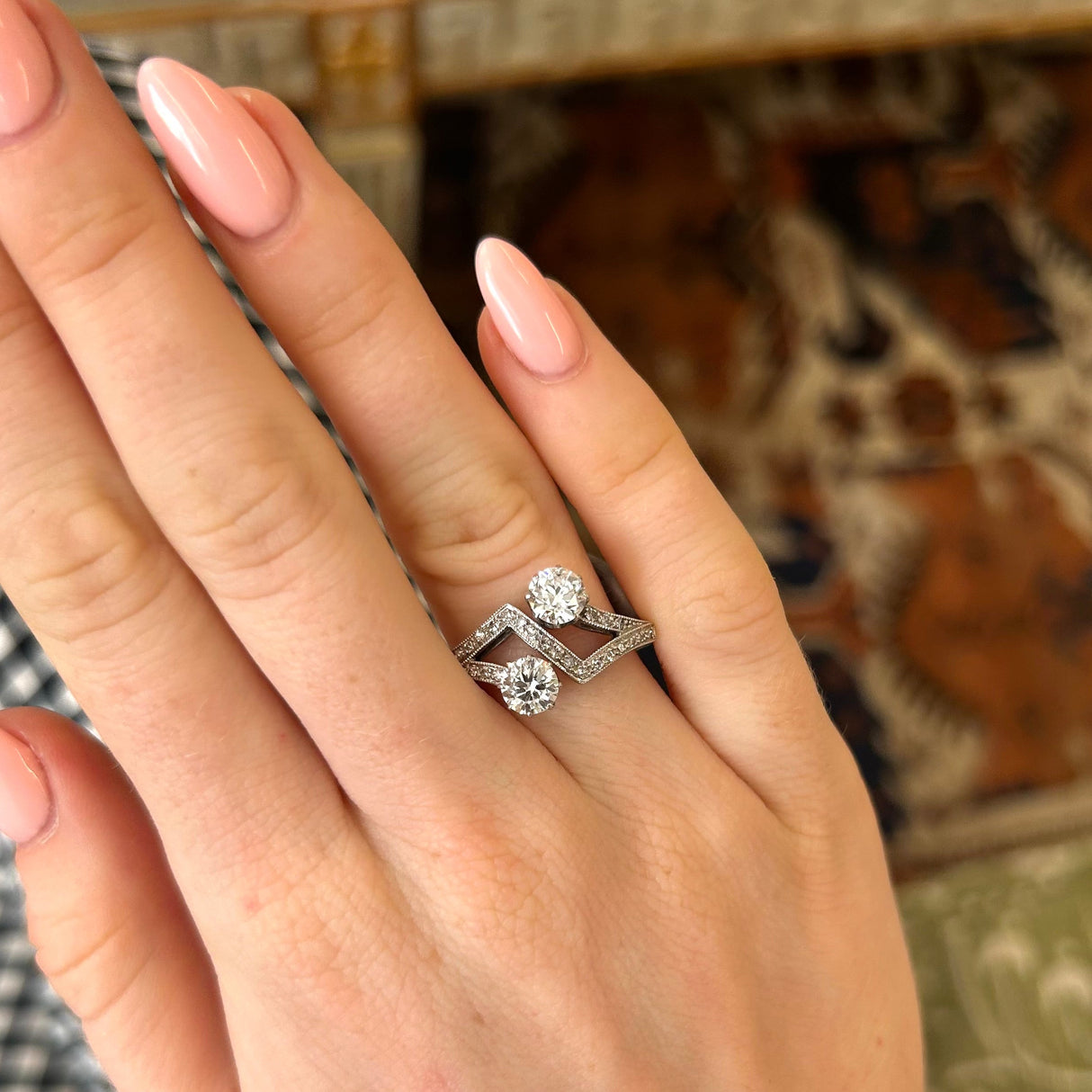 A close-up of a hand with pink manicured nails wearing a platinum antique toi et moi ring with two large diamonds set against a blurred indoor background.