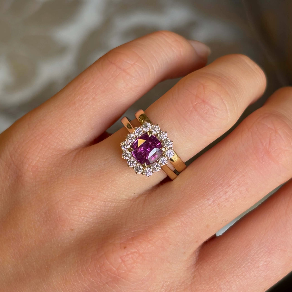 Square-cut pink sapphire and diamond cluster engagement ring and wedding band stack worn on the ring finger of the left hand close up.