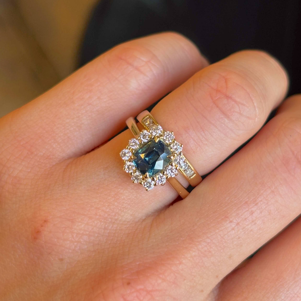 Teal sapphire and diamond cluster engagement ring paired with a wedding band on ring finger of left hand to show scale and proportions.