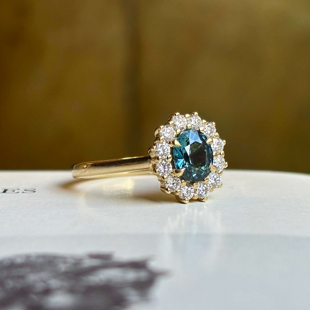 Teal sapphire and diamond yellow gold cluster engagement ring angled on light background.