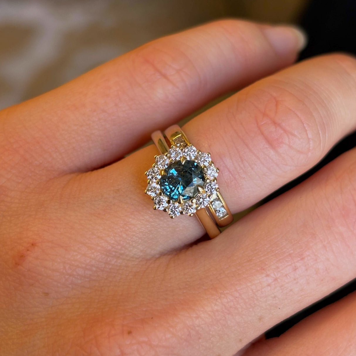 Teal sapphire and diamond cluster engagement ring worn with wedding band on hand close up