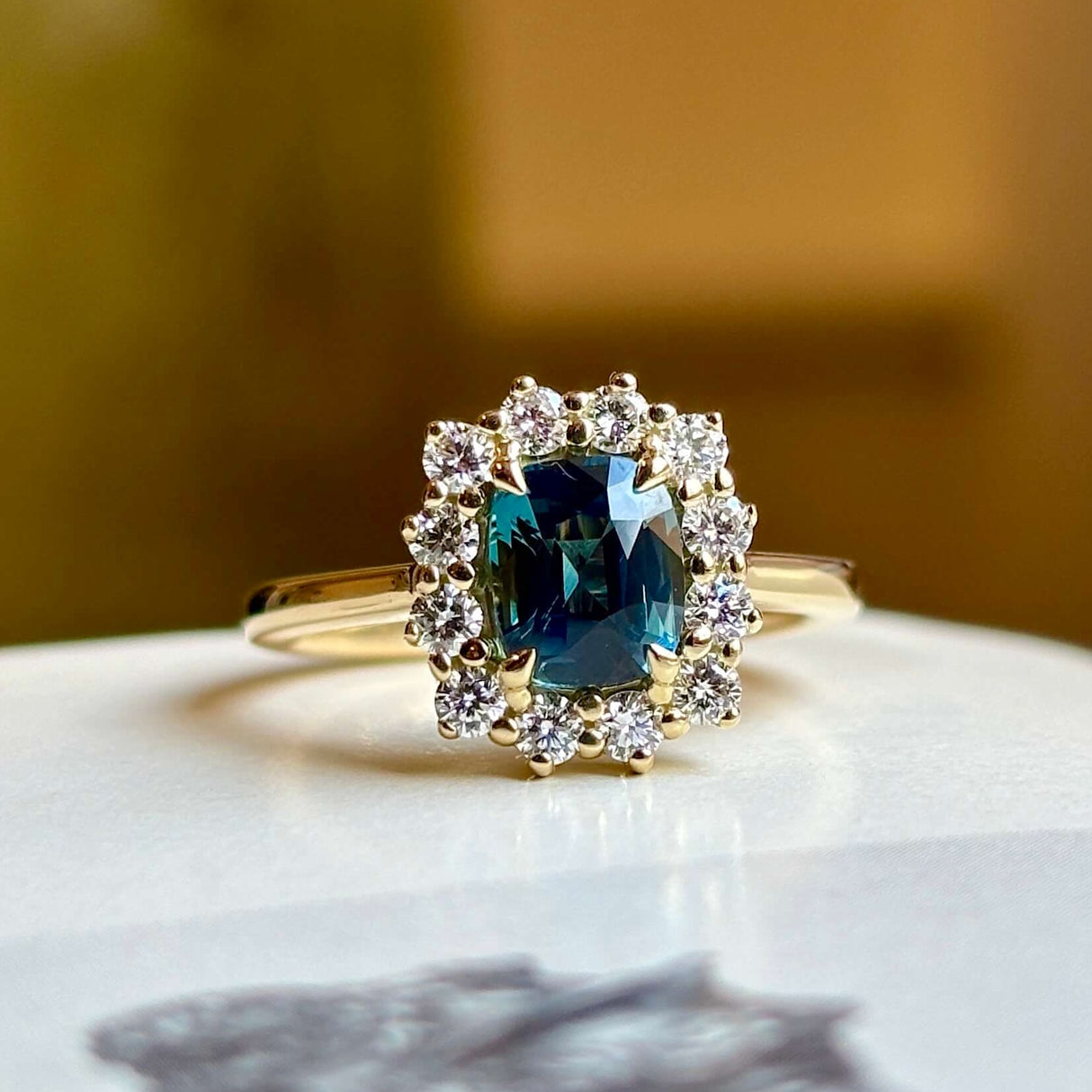 Teal sapphire and diamond cluster ring in yellow gold close-up on blurred background.