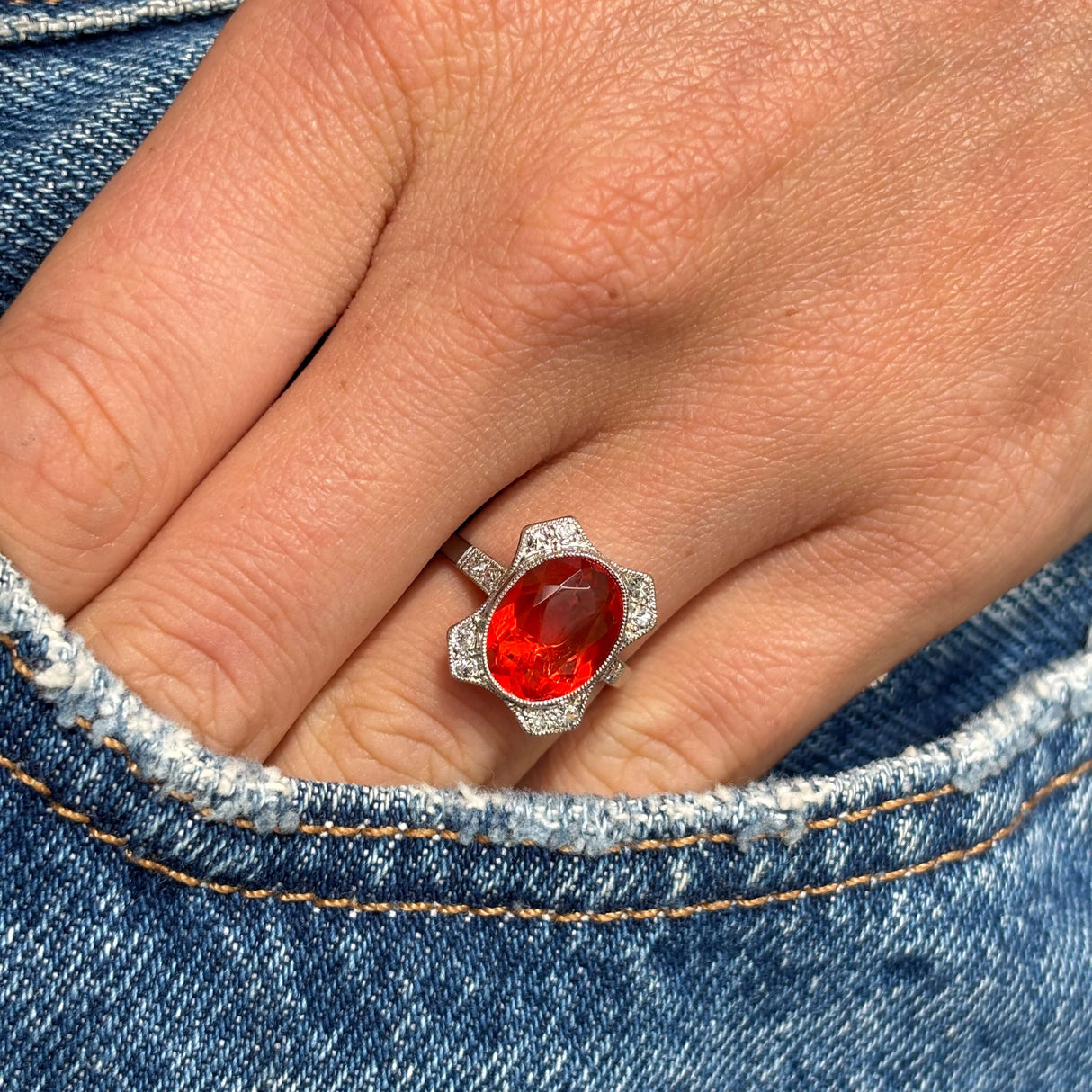 Antique Art Deco fire opal and diamond cluster ring with a vibrant fire opal centre stone and sparkling diamond halo, worn on a hand with a denim background.