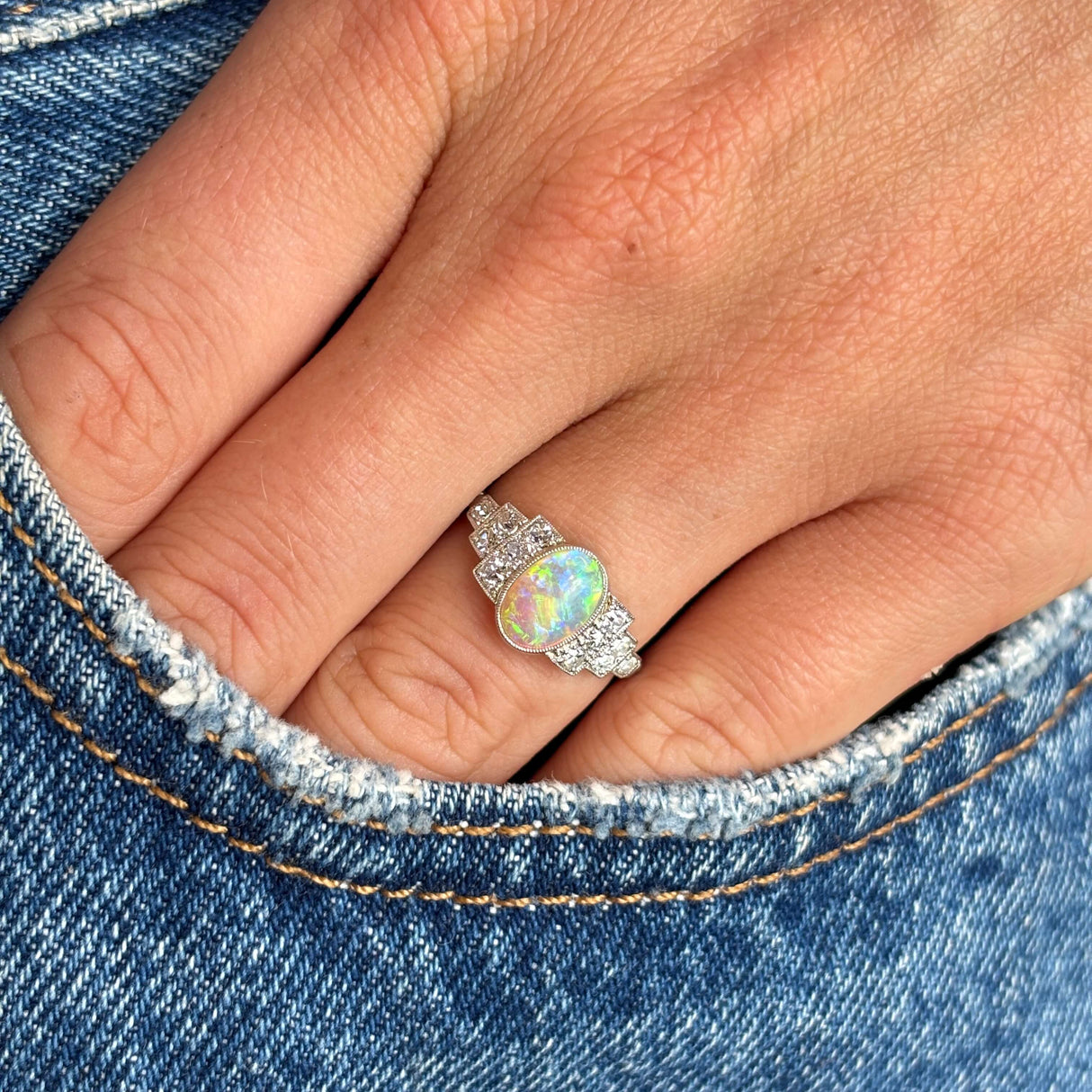 Antique Art Deco opal and diamond cluster ring photographed on hand against denim, highlighting the iridescent opal and brilliant diamond surround.