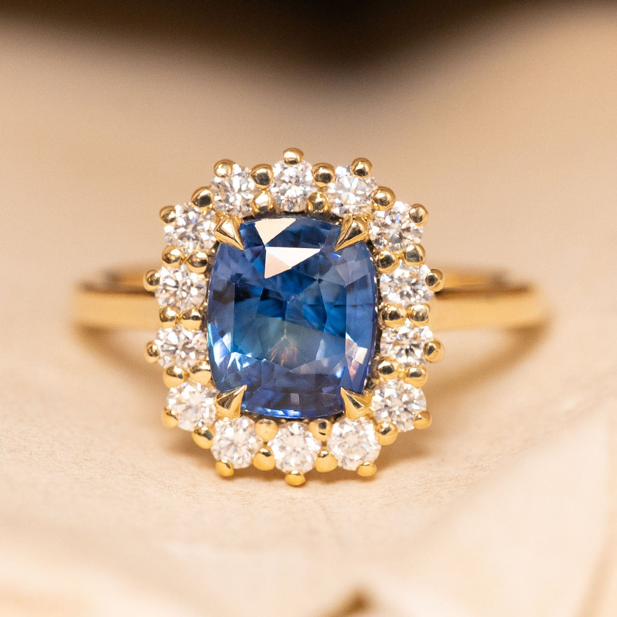 Antique-inspired blue cushion-cut sapphire and diamond cluster engagement ring featuring a richly coloured cushion-cut blue sapphire surrounded by brilliant-cut diamonds in yellow gold.