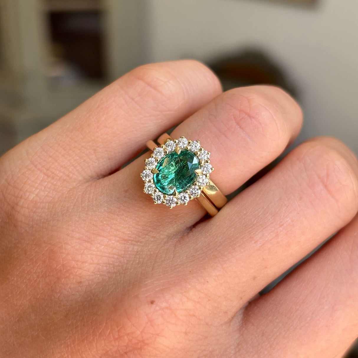 Antique-inspired yellow gold cluster ring featuring an oval-cut emerald surrounded by brilliant diamonds, photographed on hand against a softly lit vintage interior backdrop.
