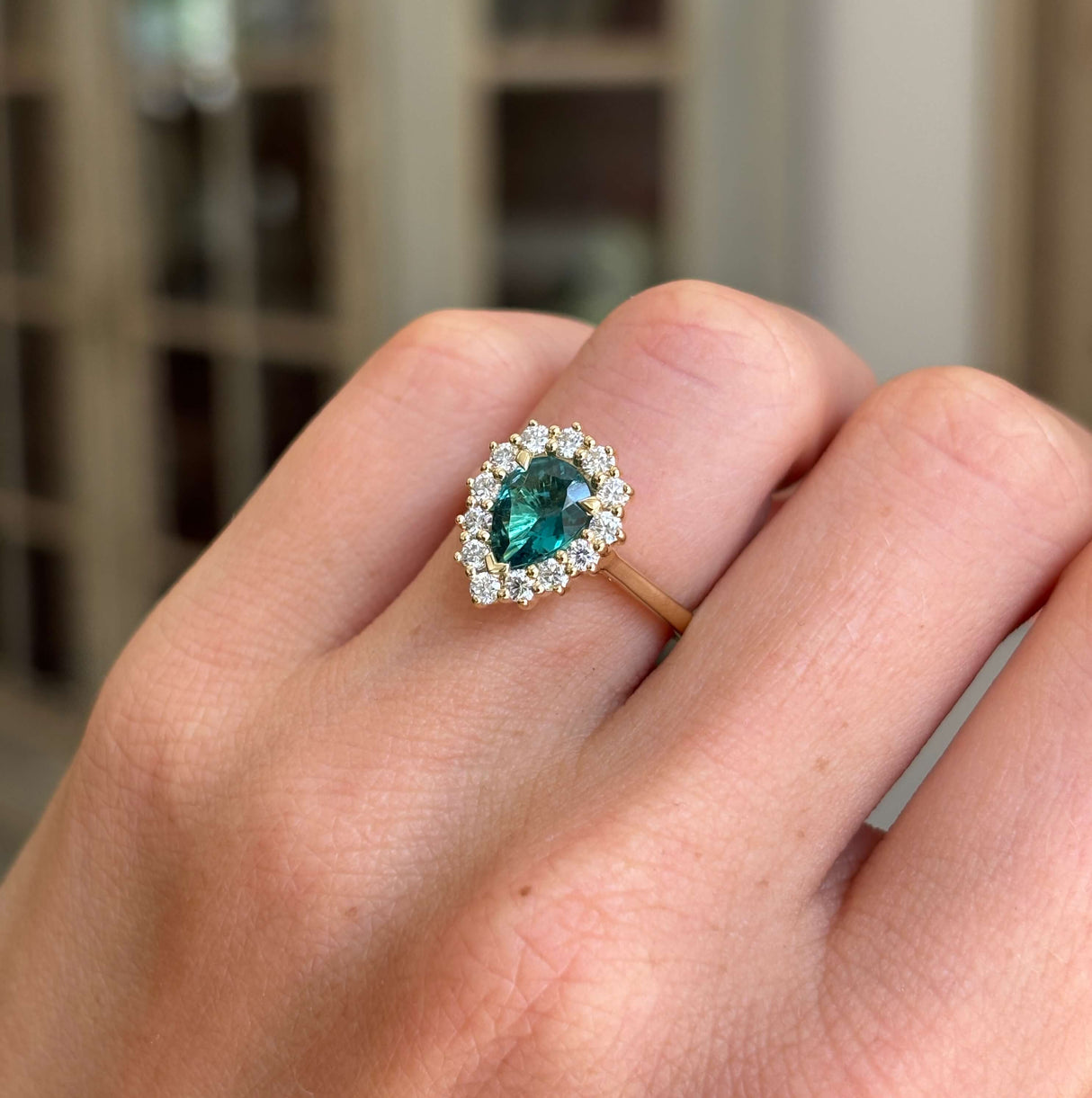 Antique-inspired pear-cut teal tourmaline and diamond cluster engagement ring featuring a pear-shaped teal tourmaline surrounded by brilliant-cut diamonds, displayed on a hand with a blurred background.