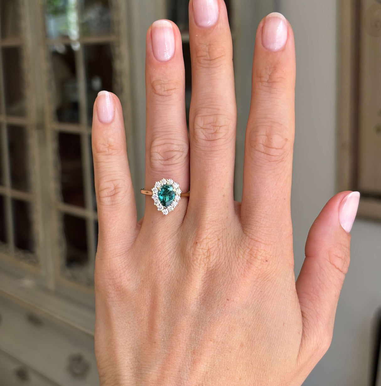 Antique-inspired pear-cut teal tourmaline and diamond cluster engagement ring with a pear-shaped teal tourmaline centre and diamond halo, worn on a hand.