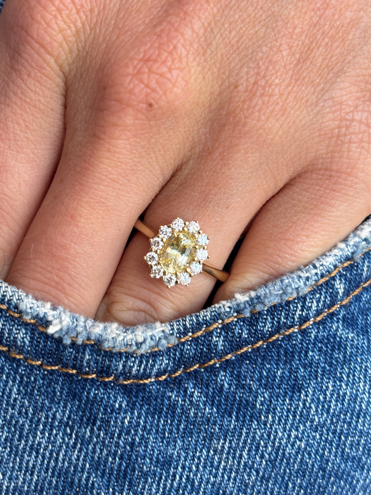 Antique-inspired yellow sapphire and diamond cluster engagement ring with an oval yellow sapphire centre stone and sparkling diamond halo, worn on a hand against a denim background