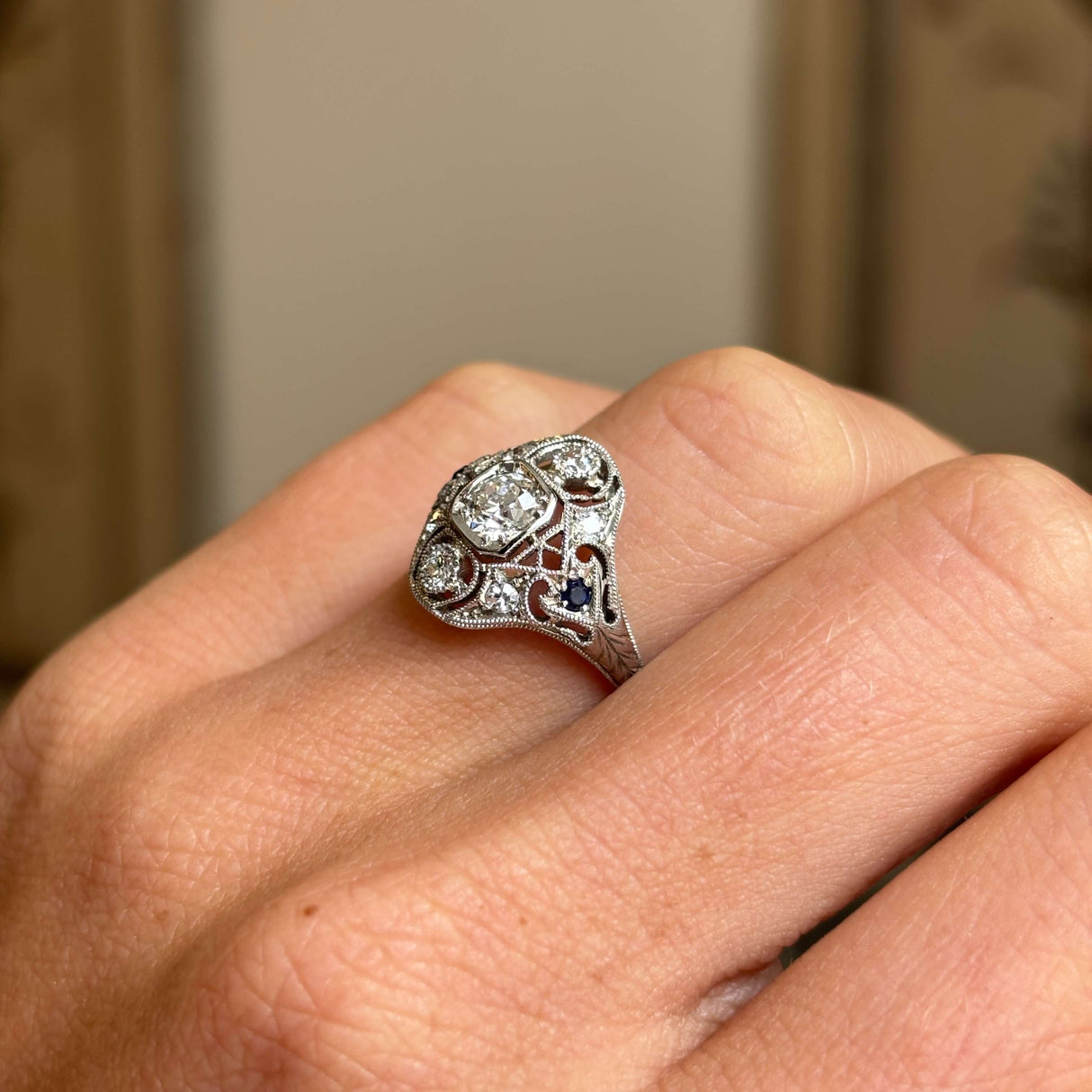Antique Art Deco sapphire and diamond cluster ring in platinum worn on hand, featuring a vivid blue sapphire surrounded by sparkling old-cut diamonds, showcasing elegant 1920s craftsmanship against a softly lit interior backdrop.