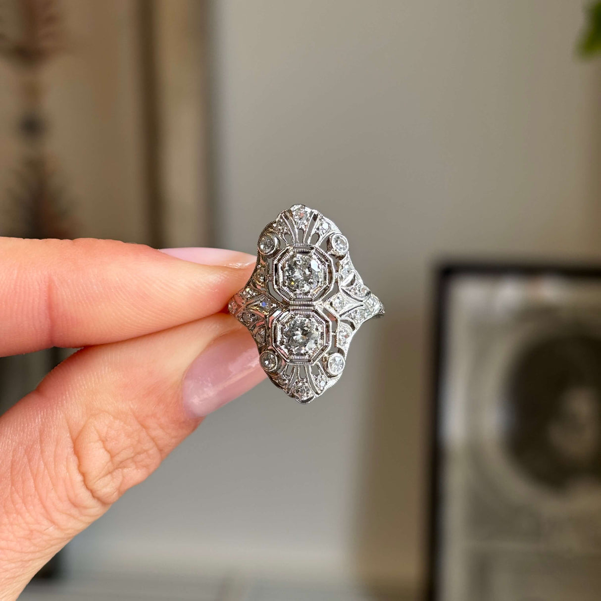 Antique Art Deco diamond cluster cocktail ring in platinum, featuring an intricate filigree setting and old-cut diamonds, held between fingers against a softly blurred vintage interior background.