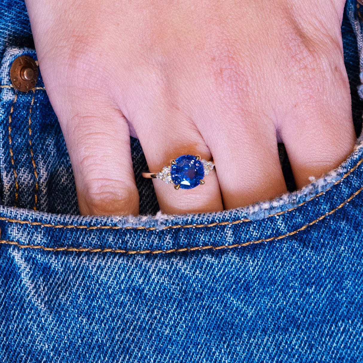 Round blue sapphire and diamond cluster tre-foil engagement ring worn on hand with denim pocket styling.
