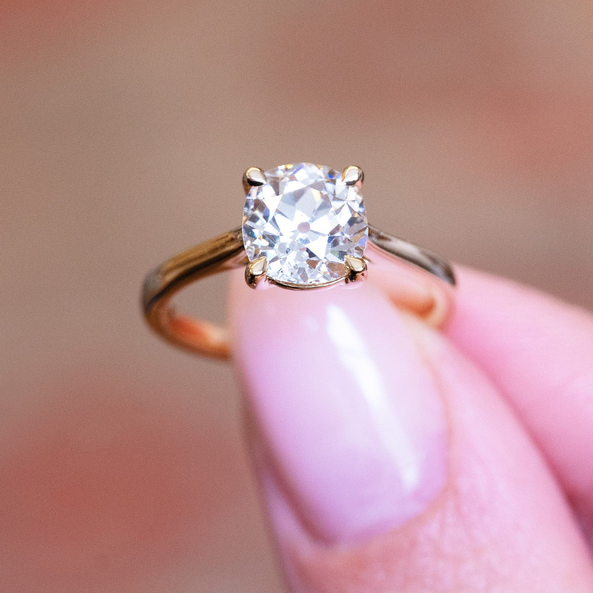 Cushion old-mine cut diamond solitaire engagement ring held between fingers with gold band.