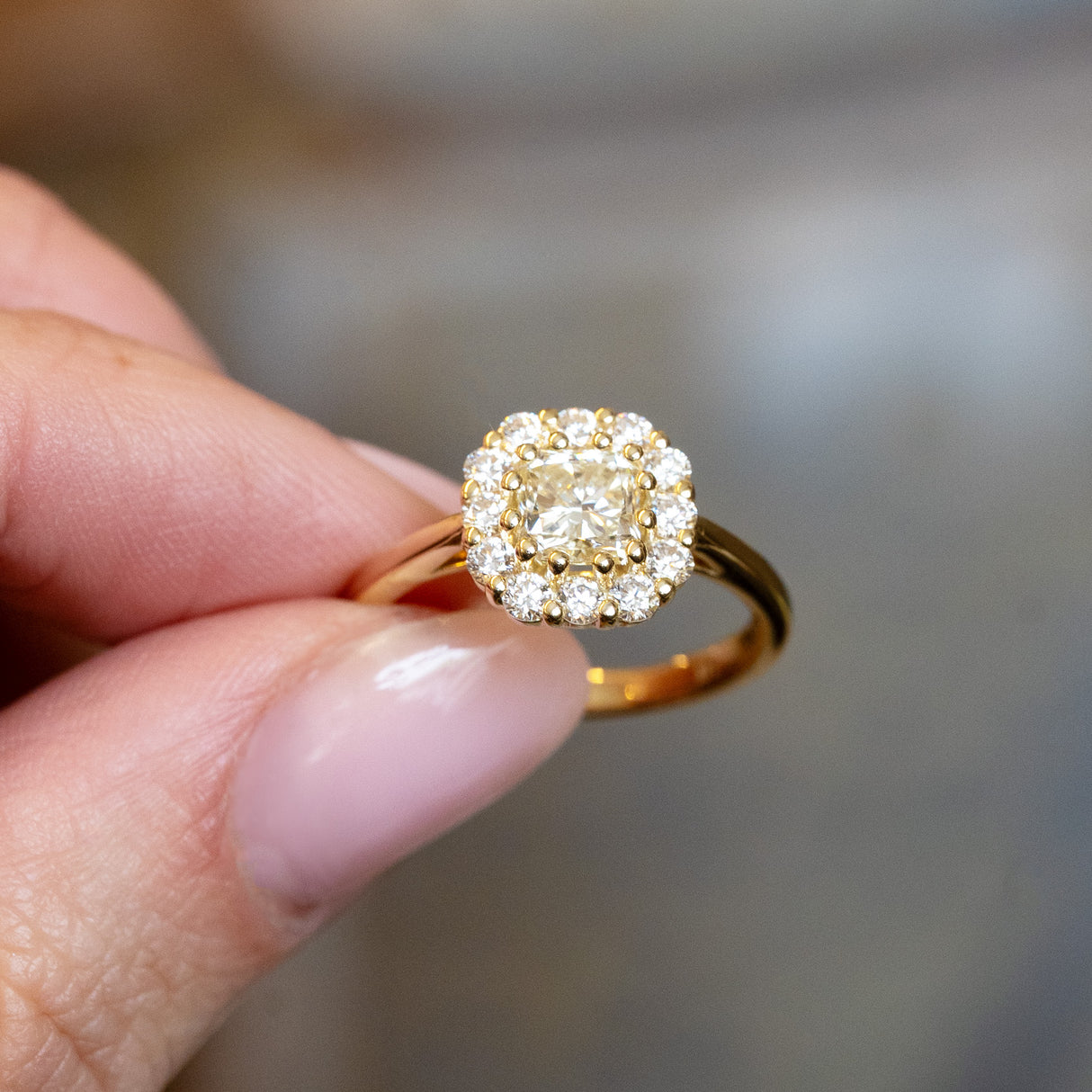 Diamond cluster engagement ring with brilliant-cut centre diamond and yellow gold band held between fingers.