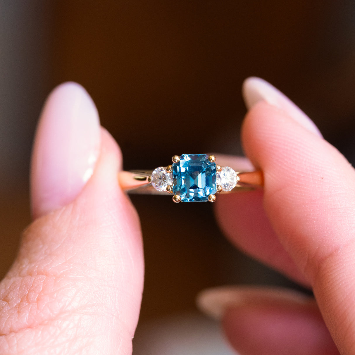 Teal sapphire and diamond three-stone engagement ring with emerald-cut teal sapphire centre stone and gold band.